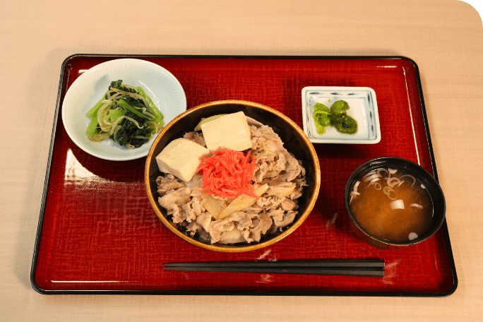 この画像も日替わり定食の写真です。お盆の上におかずが1品とお肉が乗ったどんぶり、味噌汁と漬物が乗せられています。