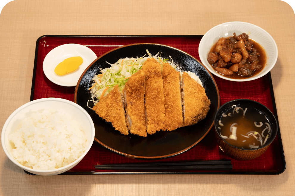 とんかつ定食の写真です。お盆の上にとんかつとキャベツが乗ったお皿があります。その周りにご飯、味噌汁、おかず、漬物がそれぞれ盛り付けられたお皿や茶碗があります。