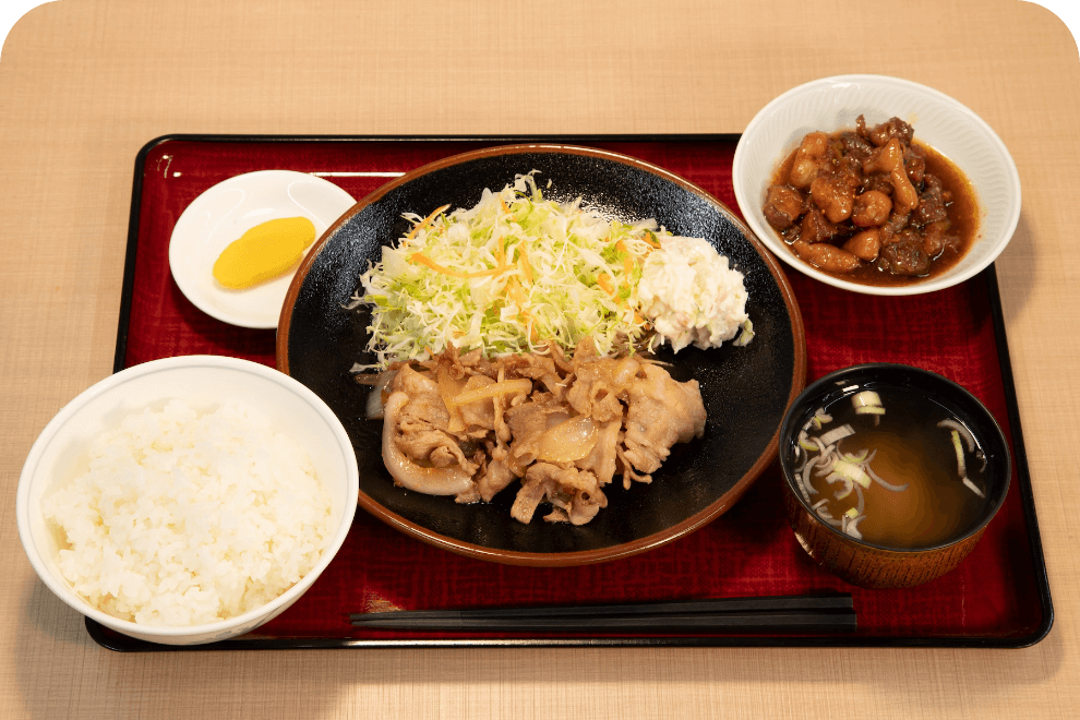 生姜焼き定食の写真です。お盆の上に生姜焼きとキャベツが乗ったお皿があります。その周りにご飯、味噌汁、おかず、漬物がそれぞれ盛り付けられたお皿や茶碗があります。